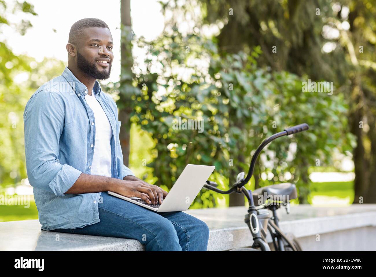 Uomo africano pensoso seduto nel parco con computer portatile, scrittura saggio all'aperto Foto Stock