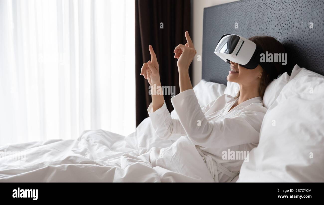 Giovane donna sorridente che indossa occhiali per realtà virtuale, dispositivo di test a letto. Foto Stock