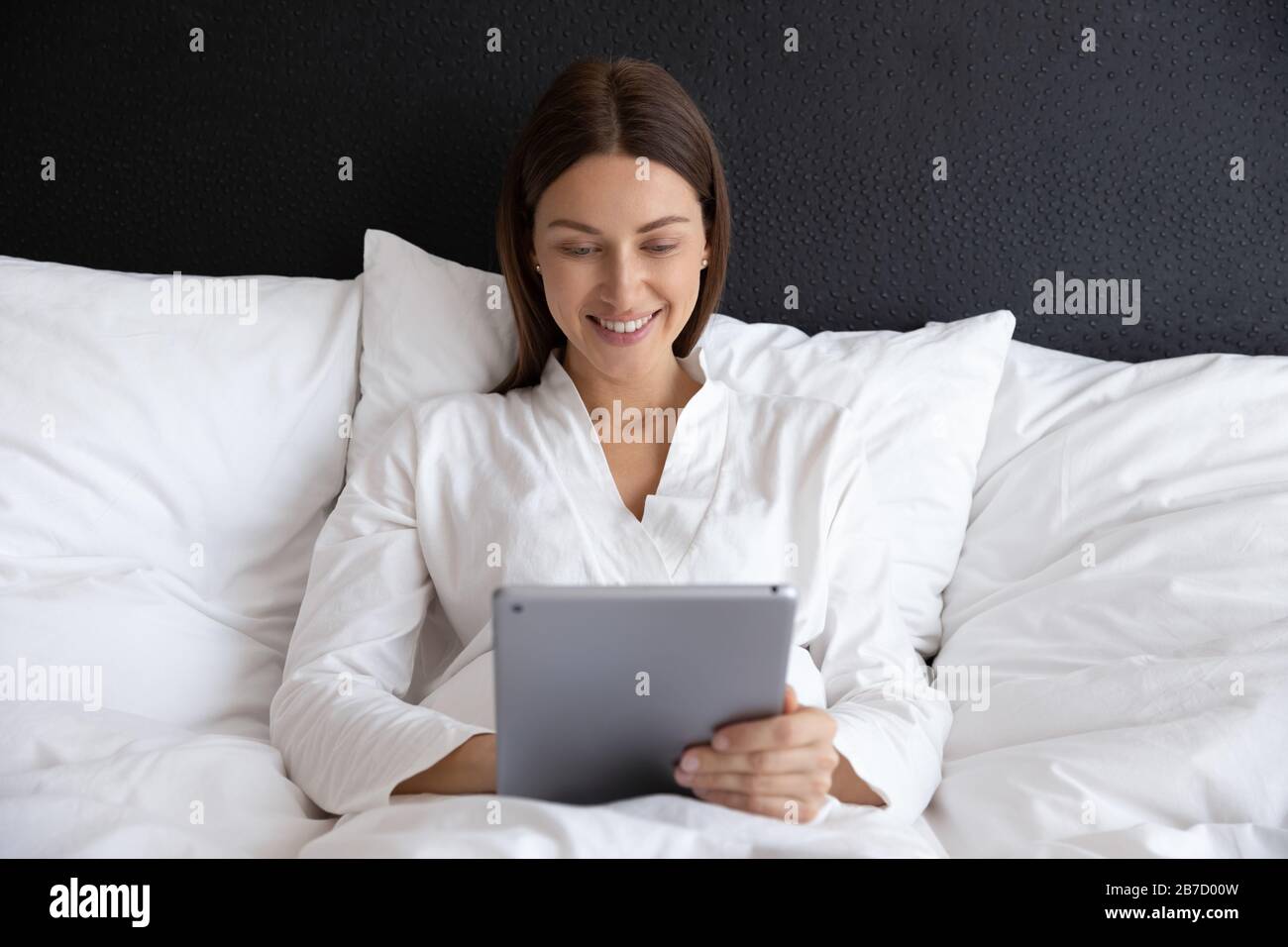 Giovane donna sorridente sdraiata su un comodo letto, utilizzando un tablet digitale. Foto Stock