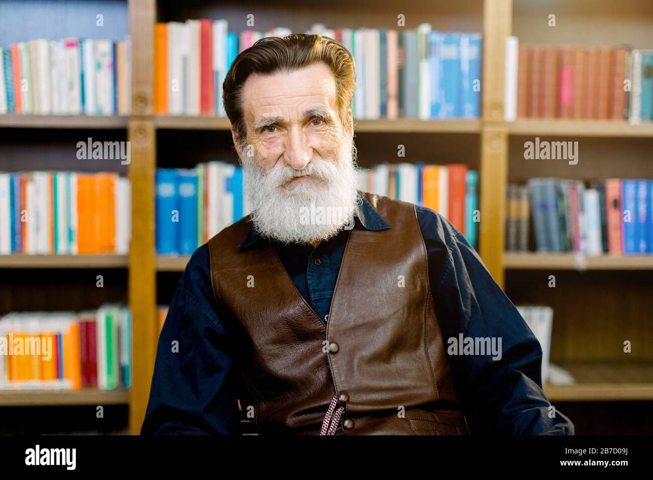 Ritratto di anziano barbuto uomo, bibliotecario o professore accademico, seduto sullo sfondo di librerie e scaffali in biblioteca o libro negozio di mercato Foto Stock
