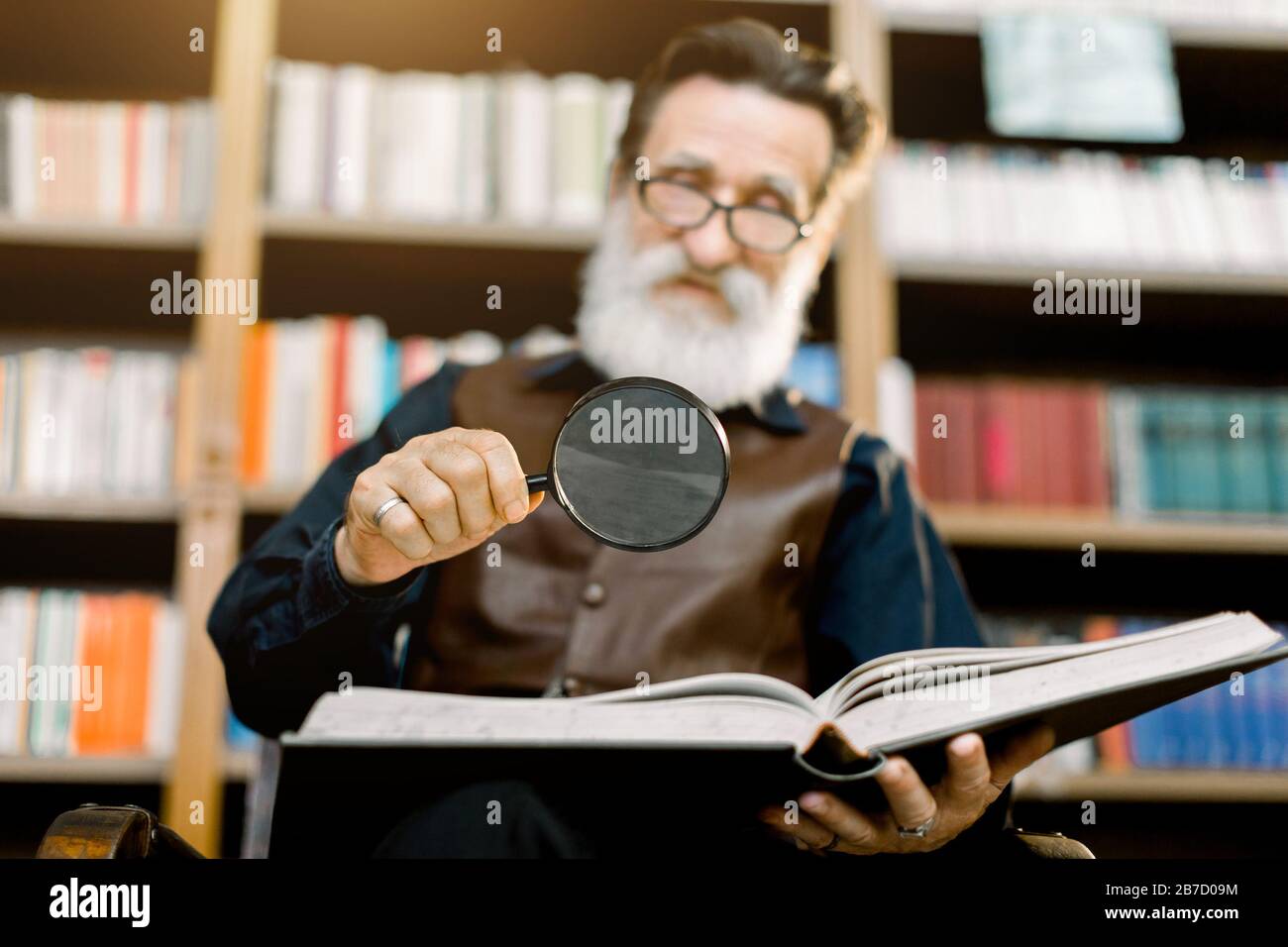 Bell'uomo anziano, barbuto, bibliotecario o professore sorridente, in biblioteca, seduto sullo sfondo di librerie, con lente d'ingrandimento e Foto Stock