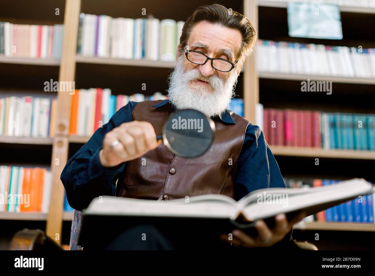 Bell'uomo pensoso anziano barbuto, bibliotecario o professore, in biblioteca, seduto sullo sfondo di librerie, con lente d'ingrandimento e Foto Stock