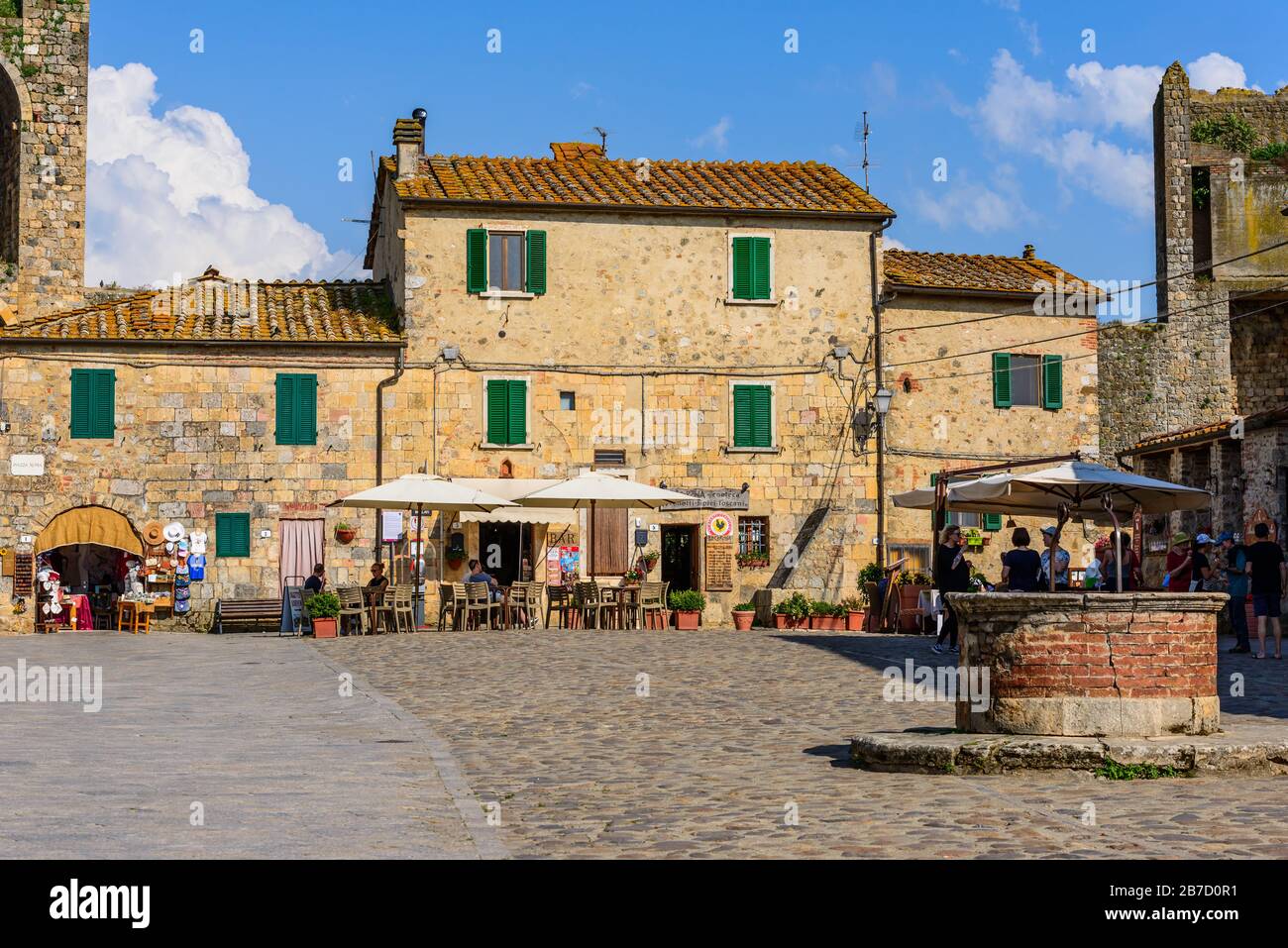 Un pozzo nel mezzo di Piazza Roma, Monteriggioni, Toscana, Italia, circondato da edifici storici, alcuni negozi, ristoranti e gelaterie Foto Stock