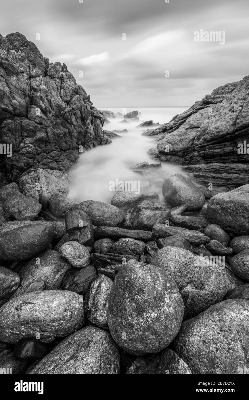 Una fotografia verticale del paesaggio del tramonto in bianco e nero di onde nebbie che si schiantano sulle rocce dalla costa meridionale in Sudafrica Foto Stock