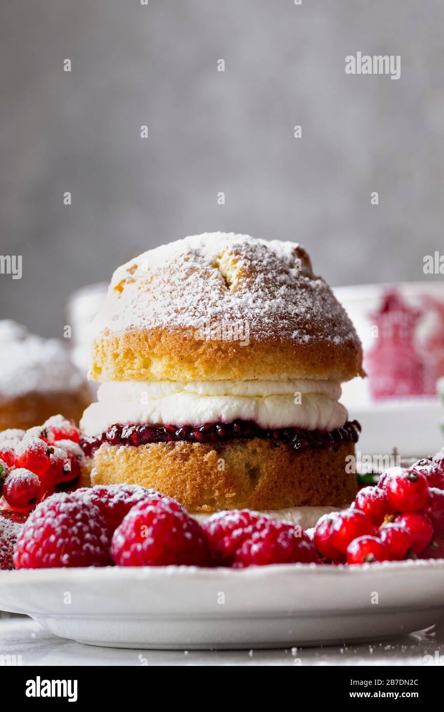 Torta Victoria Sponge con marmellata di lamponi e panna montata, generosamente cosparsa di zucchero in polvere, servita con frutta. Foto Stock