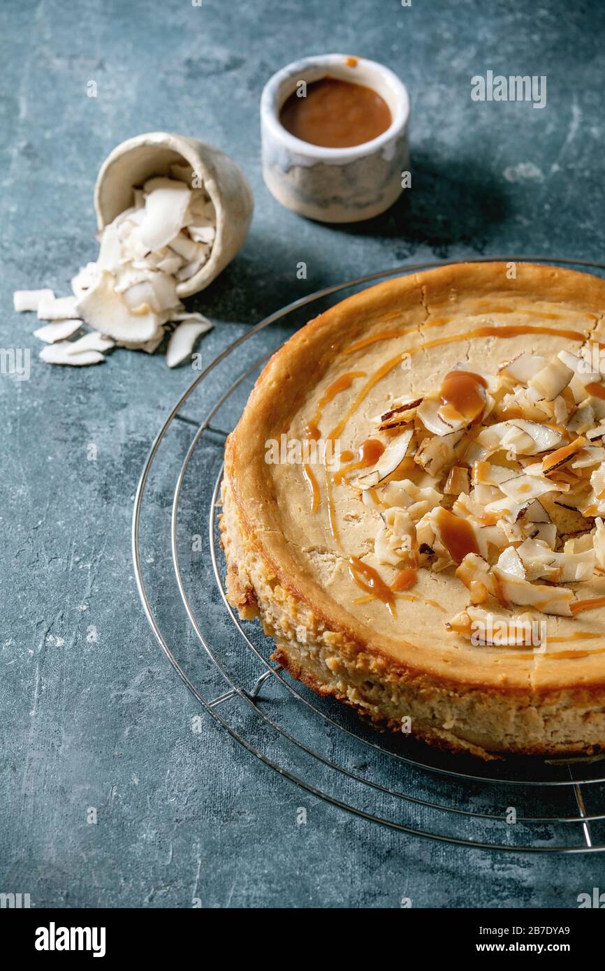 Torta di cheesecake fatta in casa con zucchero di canna di cocco su griglia di raffreddamento con salsa al caramello in ciotola di ceramica su sfondo di texture blu. Foto Stock