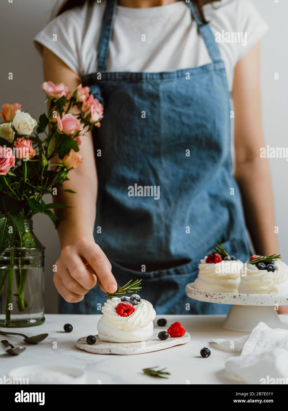 Donna in un grembiule tiene rosmarino con le dita per decorare mini Pavlova torta con frutti di bosco freschi. Composizione verticale con piano in marmo bianco an Foto Stock