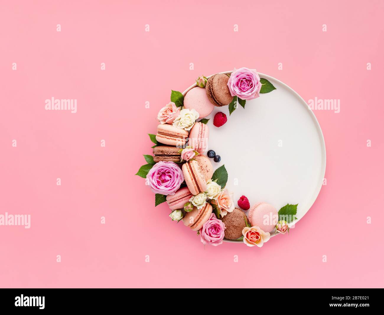 Bella composizione con macaroni francesi, fiori e bacche su piatto artigianale su sfondo rosa. Bella disposizione creativa di macaron pastello o m Foto Stock