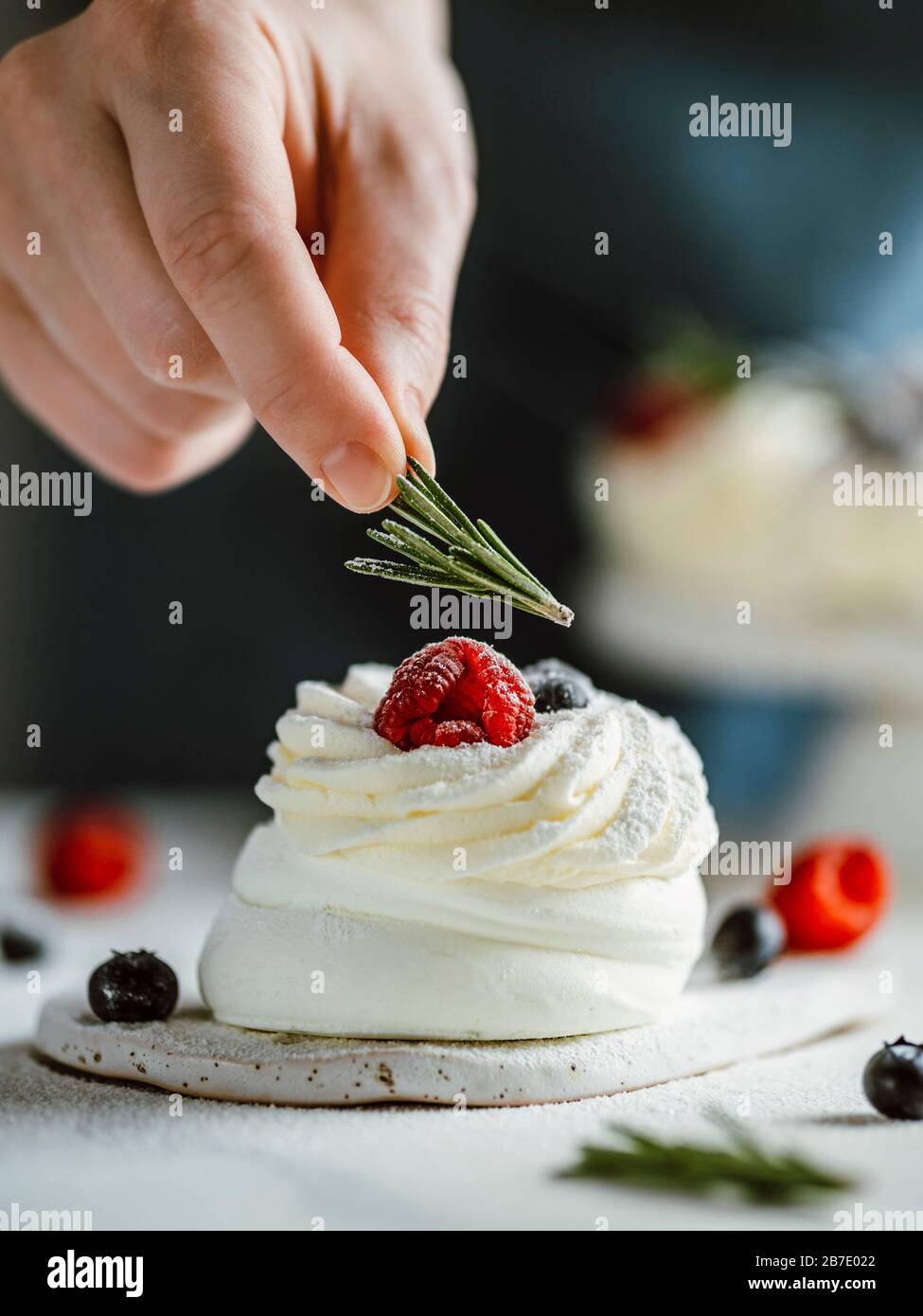 Le dita femmina trattengono il rosmarino per decorare la mini torta Pavlova con frutti di bosco freschi. Primo piano di composizione verticale con piano in marmo bianco e retro scuro Foto Stock