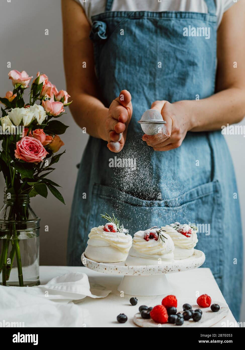 Donna in un grembiule spolga lo zucchero a velo attraverso un setaccio su mini Pavlova torte con frutti di bosco freschi. Composizione verticale con piano in marmo bianco an Foto Stock