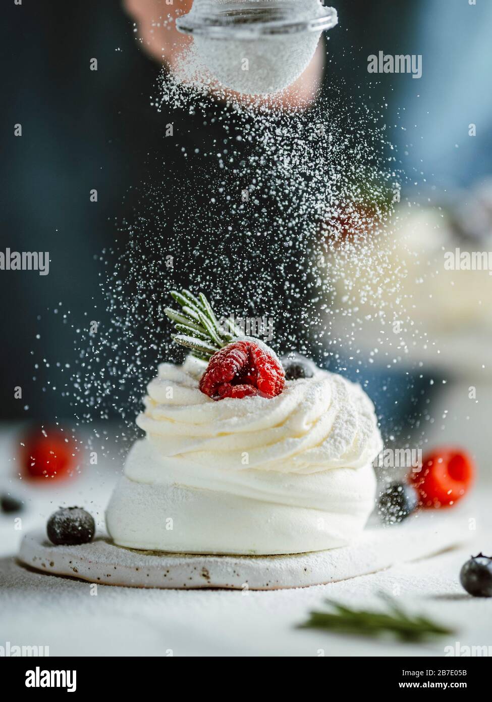 Donna spruzza lo zucchero a velo attraverso un setaccio su mini Pavlova torta decorata frutti di bosco freschi e rosmarino. Composizione verticale. Foto Stock