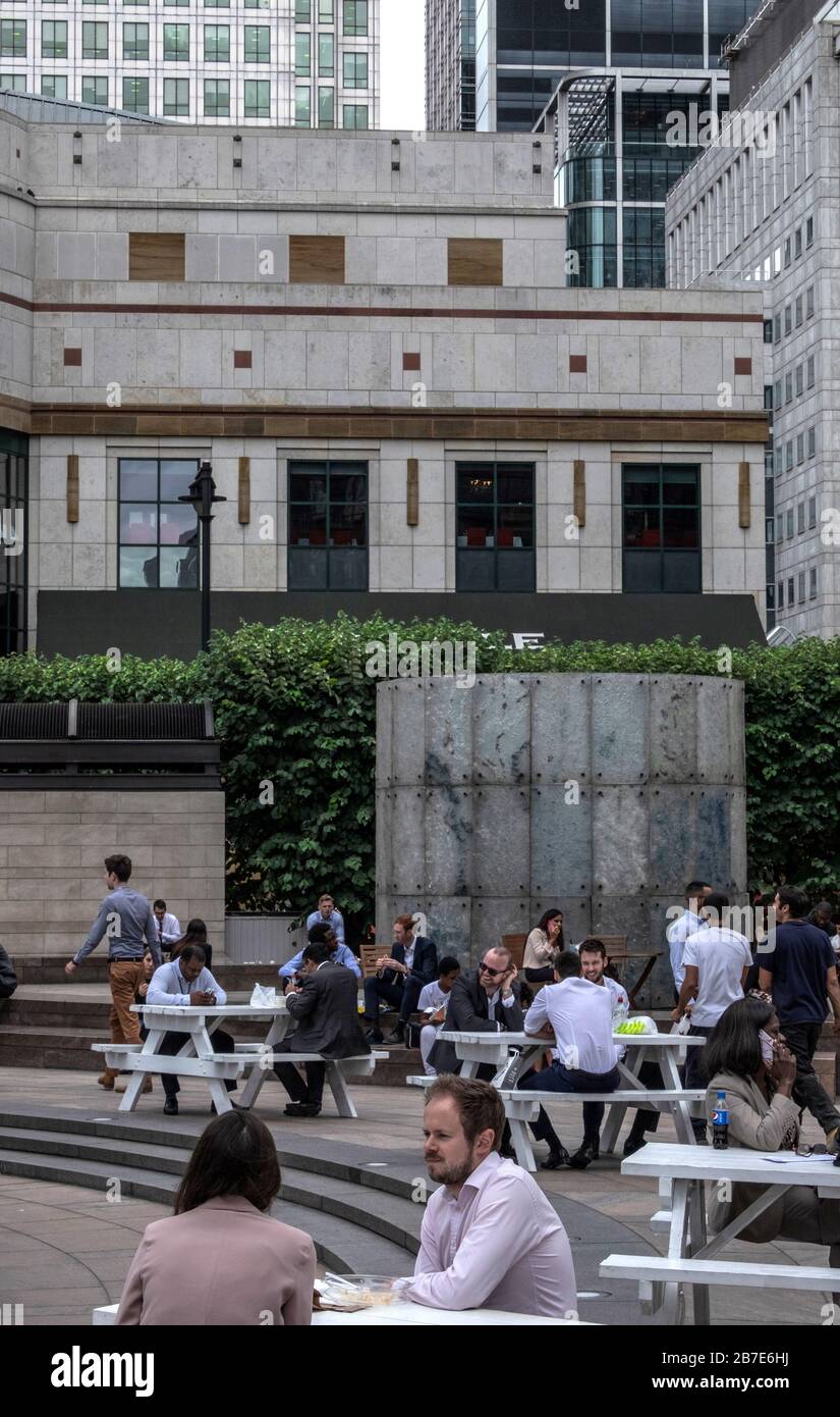 Gli impiegati di Londra mangiano il pranzo, chiacchierano & socializzano ai tavoli esterni a Cabot Square, Canary Wharf, in una giornata calda. Alti edifici di uffici sullo sfondo. Foto Stock