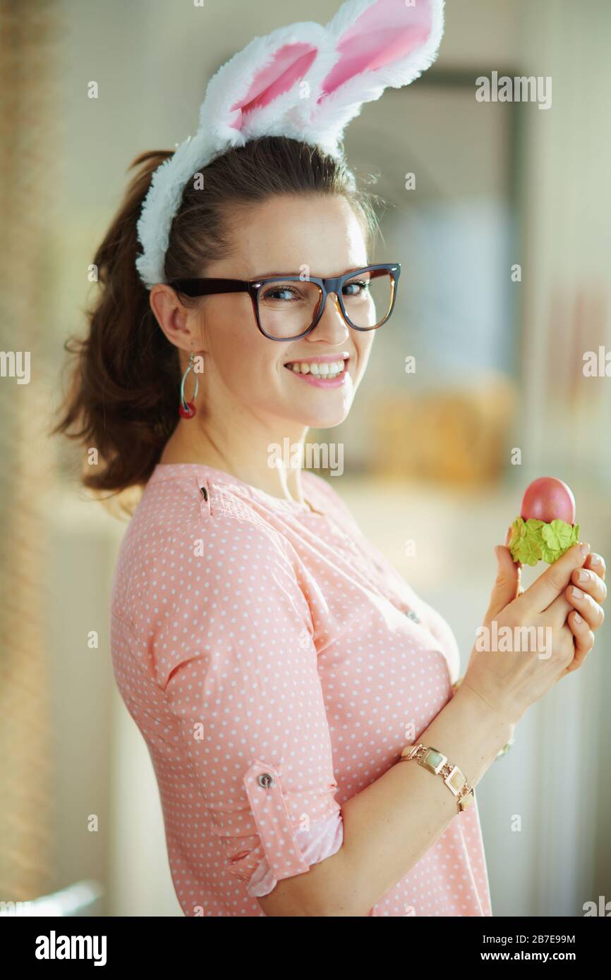 femmina moderna sorridente in una blusa rosa e orecchie di coniglietto pasquale con bicchieri che tengono uovo di pasqua rosso in una tazza di uovo verde a casa moderna nel giorno di primavera soleggiato. Foto Stock