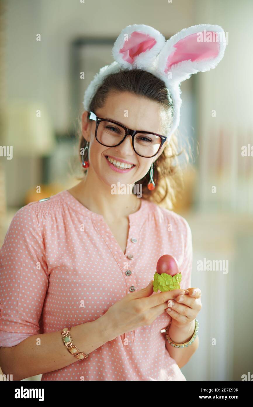 donna moderna sorridente in una blusa rosa e spighe conigliate di pasqua con bicchieri che tengono uovo di pasqua rosso in tazza di uovo verde a casa moderna in sole primavera giorno. Foto Stock