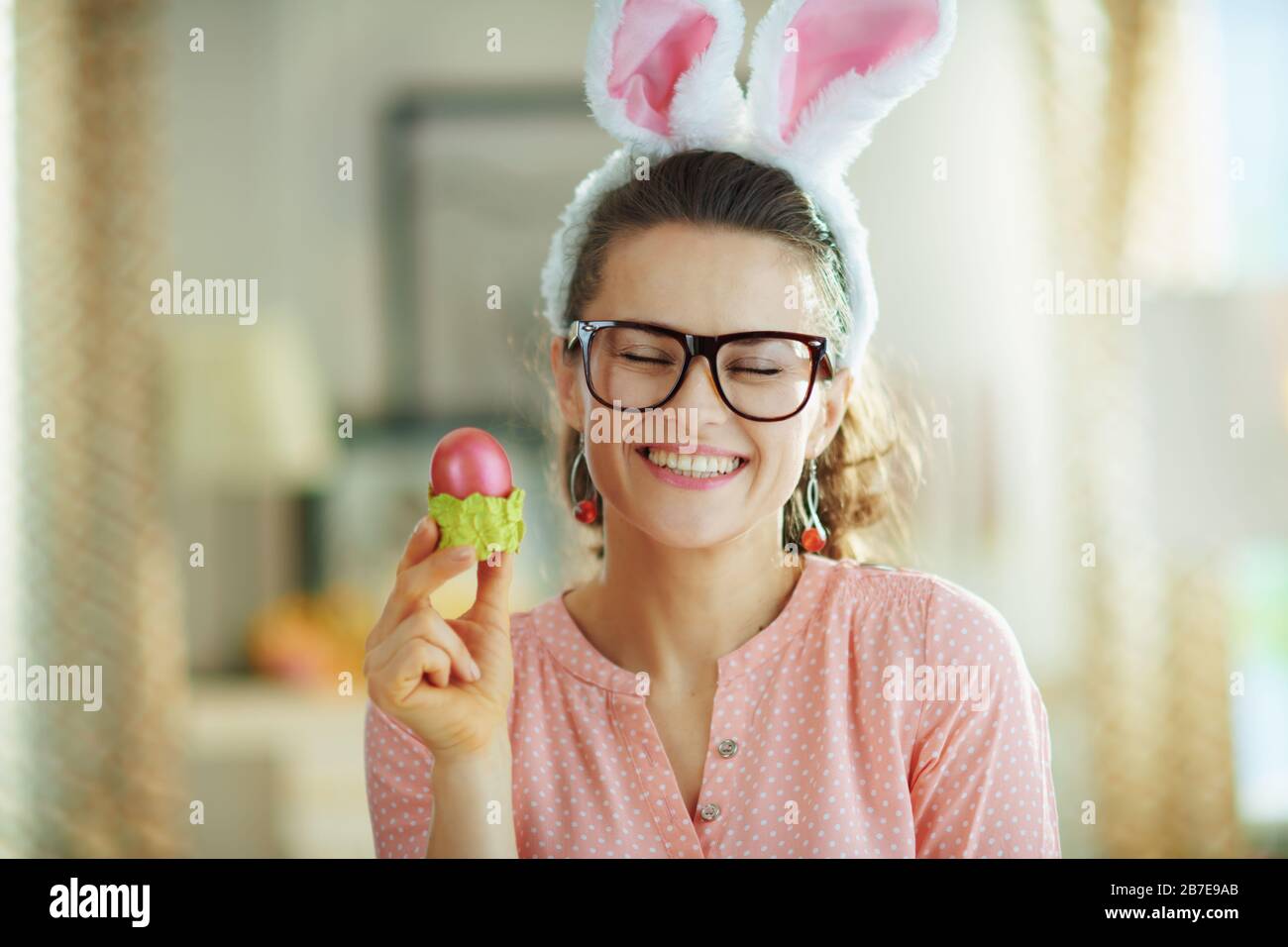 casalinga moderna sorridente in una blusa rosa e spighe conigliate di pasqua con i vetri che tengono l'uovo rosso di pasqua nella tazza verde dell'uovo alla casa moderna in da di primavera soleggiato Foto Stock