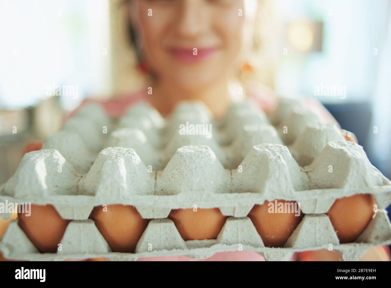 Primo piano su donna sorridente nel soggiorno in sole primavera giorno mostrando grande scatola di cartone uovo. Foto Stock