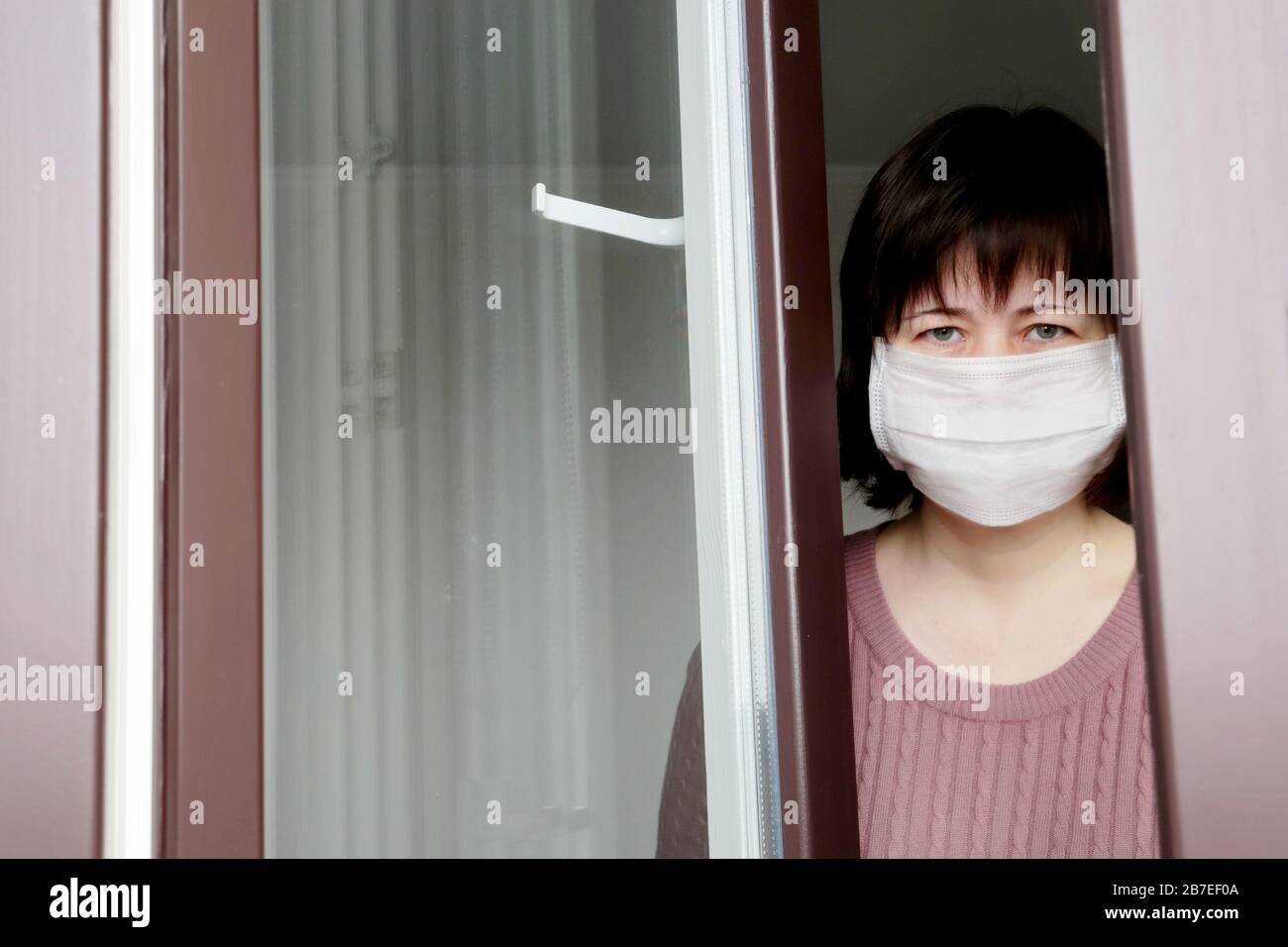 Quarantena durante l'epidemia di coronavirus COVID-19. Donna triste in una maschera medica nella finestra Foto Stock