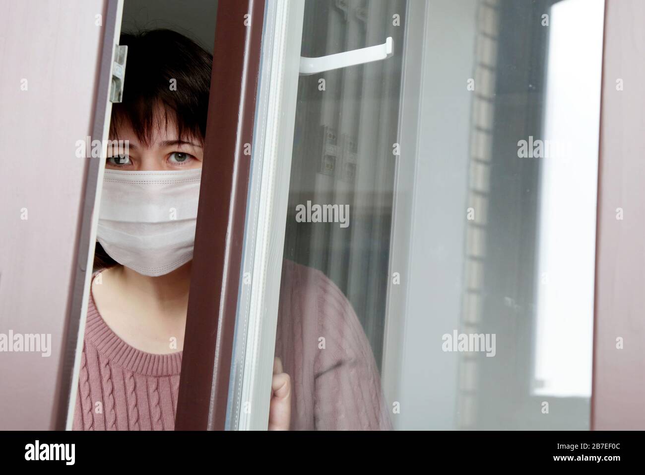 Quarantena durante l'epidemia di coronavirus COVID-19. Donna triste in una maschera medica nella finestra Foto Stock