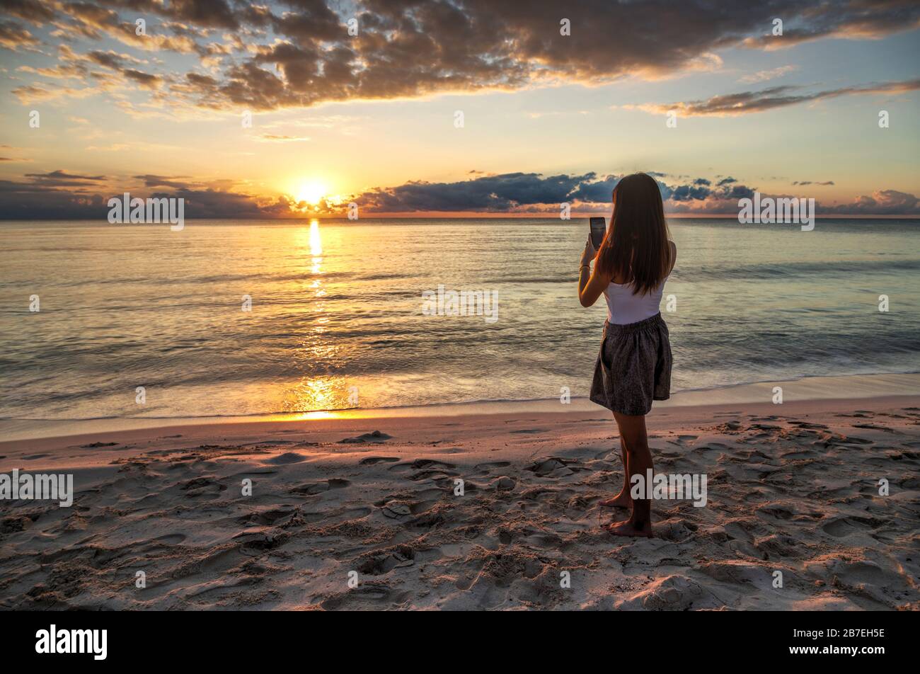 Giovane ragazza che cattura i ricordi delle vacanze di una bella alba dorata sulla spiaggia della Riviera Maya a Cancun, Messico, con il suo smartphone. Foto Stock