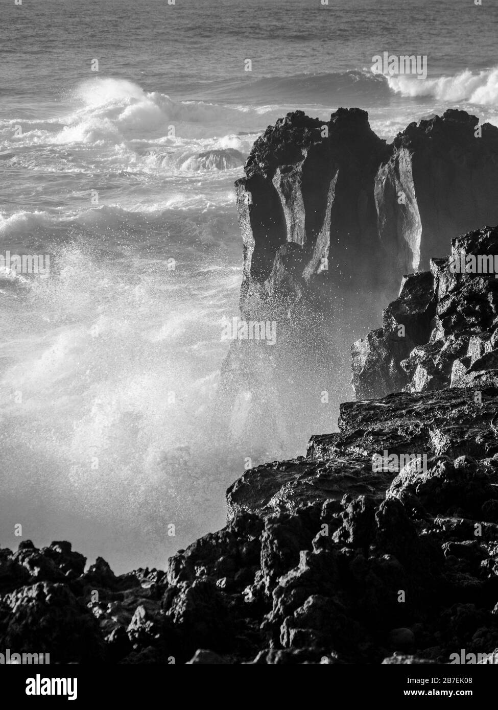 Immagine in scala di grigi di onde forti che colpiscono grandi rocce su un puntate con spruzzi d'acqua e spray Foto Stock