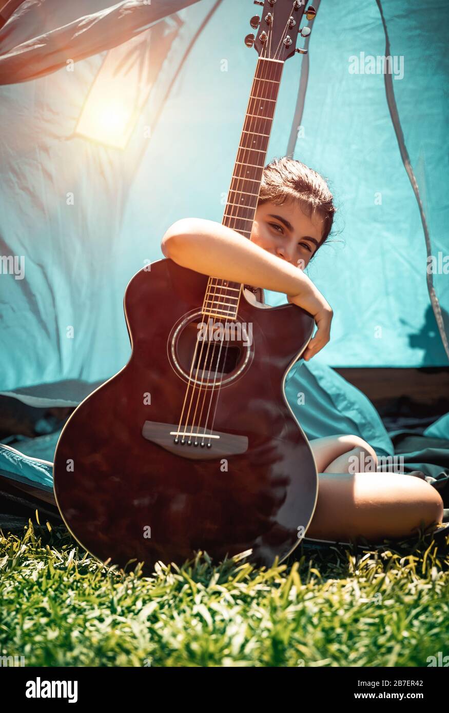 Bella ragazza adolescente seduta sulla fresca erba verde con chitarra, bambino di talento suona uno strumento musicale, godendo del tempo attivo al campo estivo Foto Stock