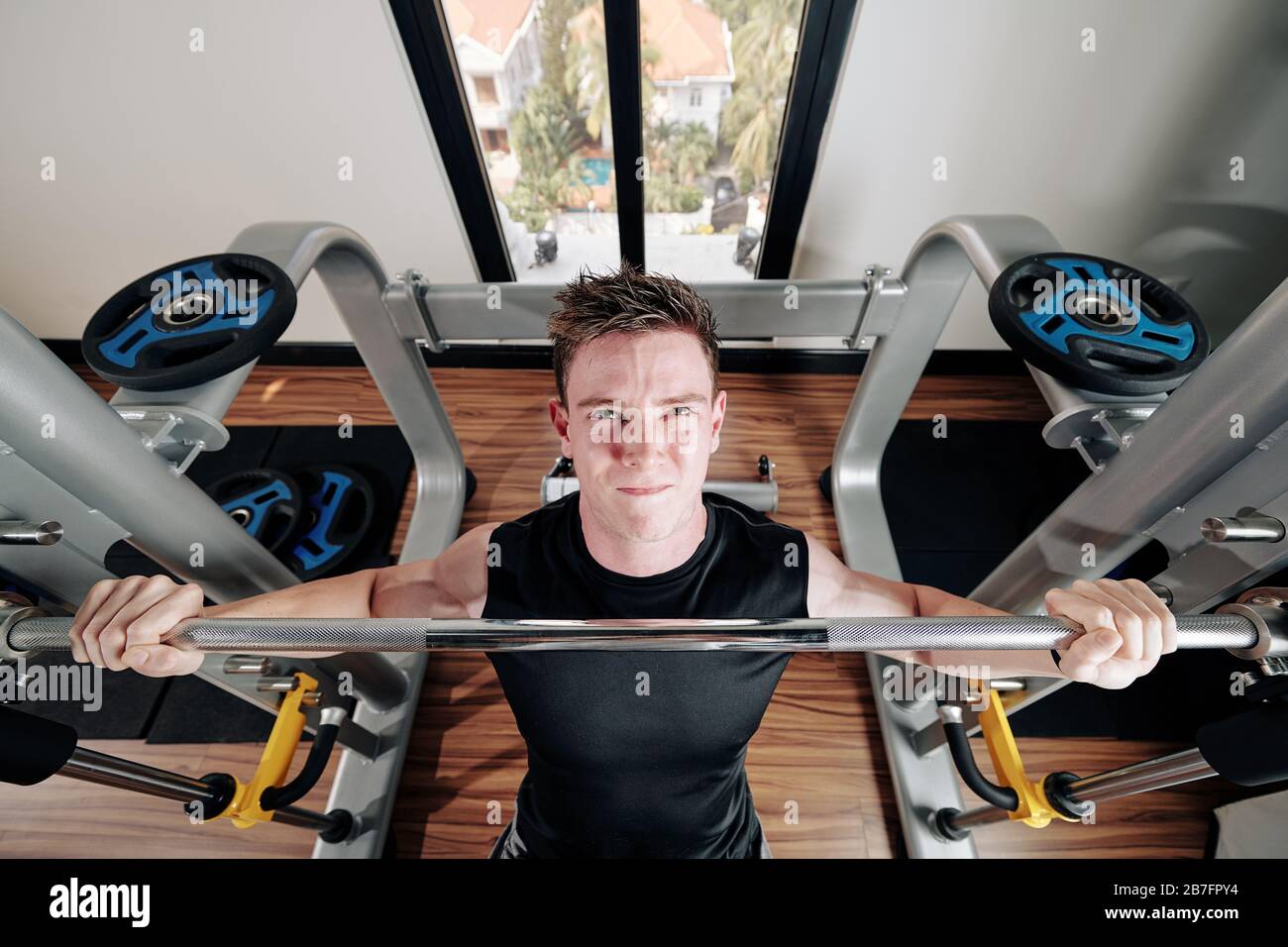 Bel giovane uomo forte che guarda in alto quando si siede su panchina sollevando pesante barbell, vista dall'alto Foto Stock