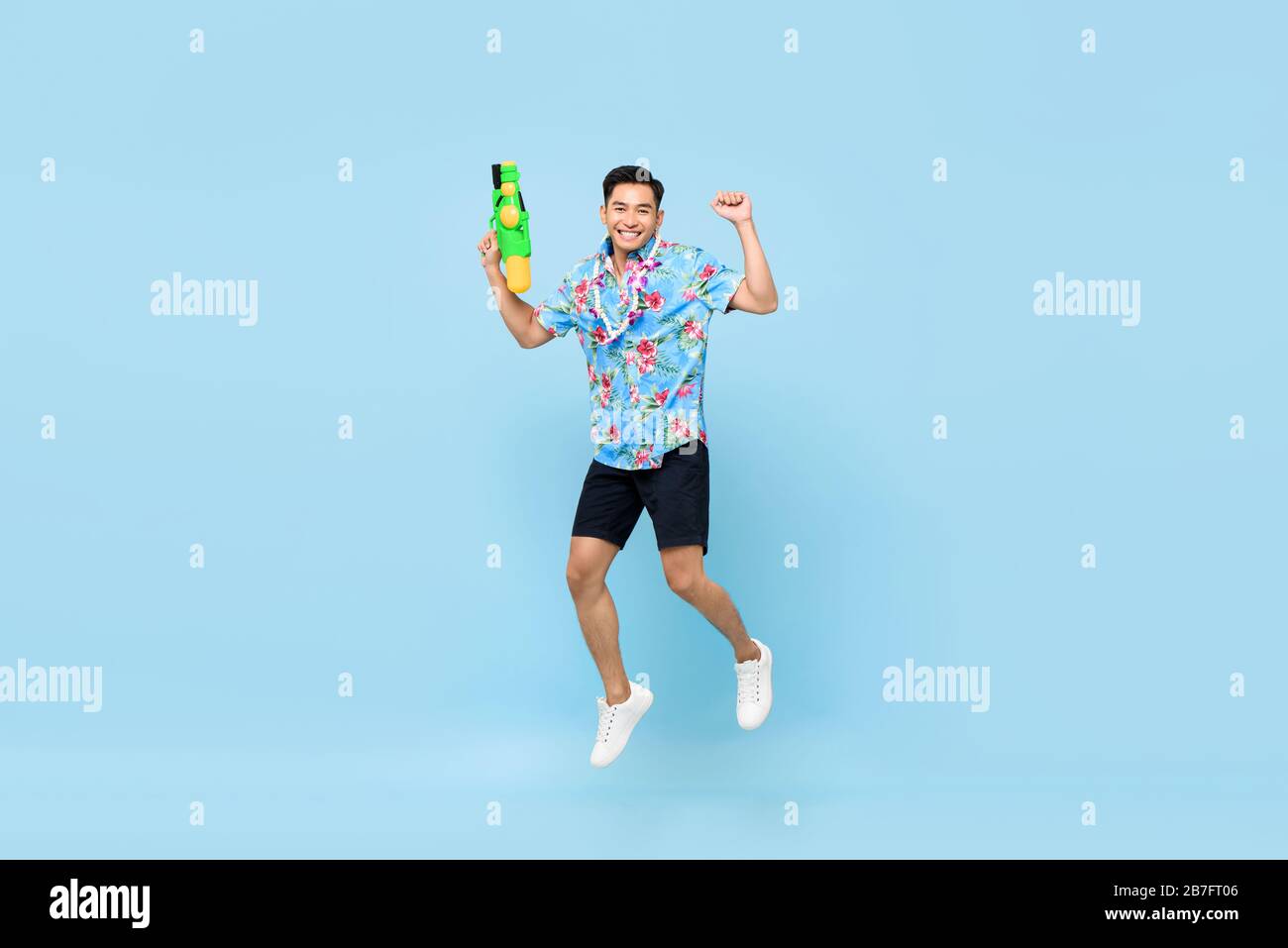 Bel giovane asiatico sorridente che gioca con pistola ad acqua e salta in studio sfondo blu per il festival Songkran in Thailandia e nel sud-est asiatico Foto Stock