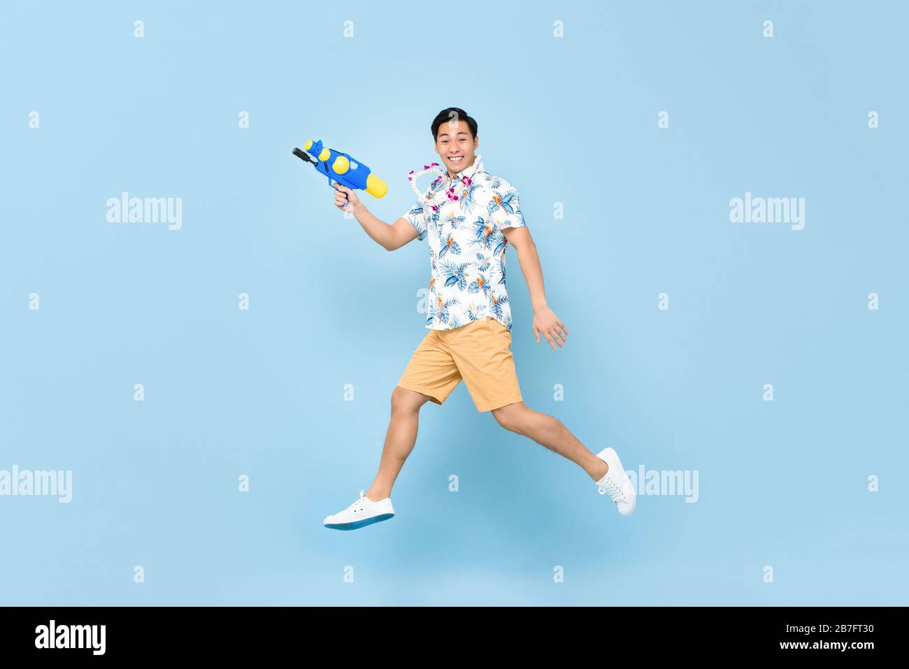 Bel giovane asiatico sorridente che gioca con pistola ad acqua e salta in studio sfondo blu per il festival Songkran in Thailandia e nel sud-est asiatico Foto Stock