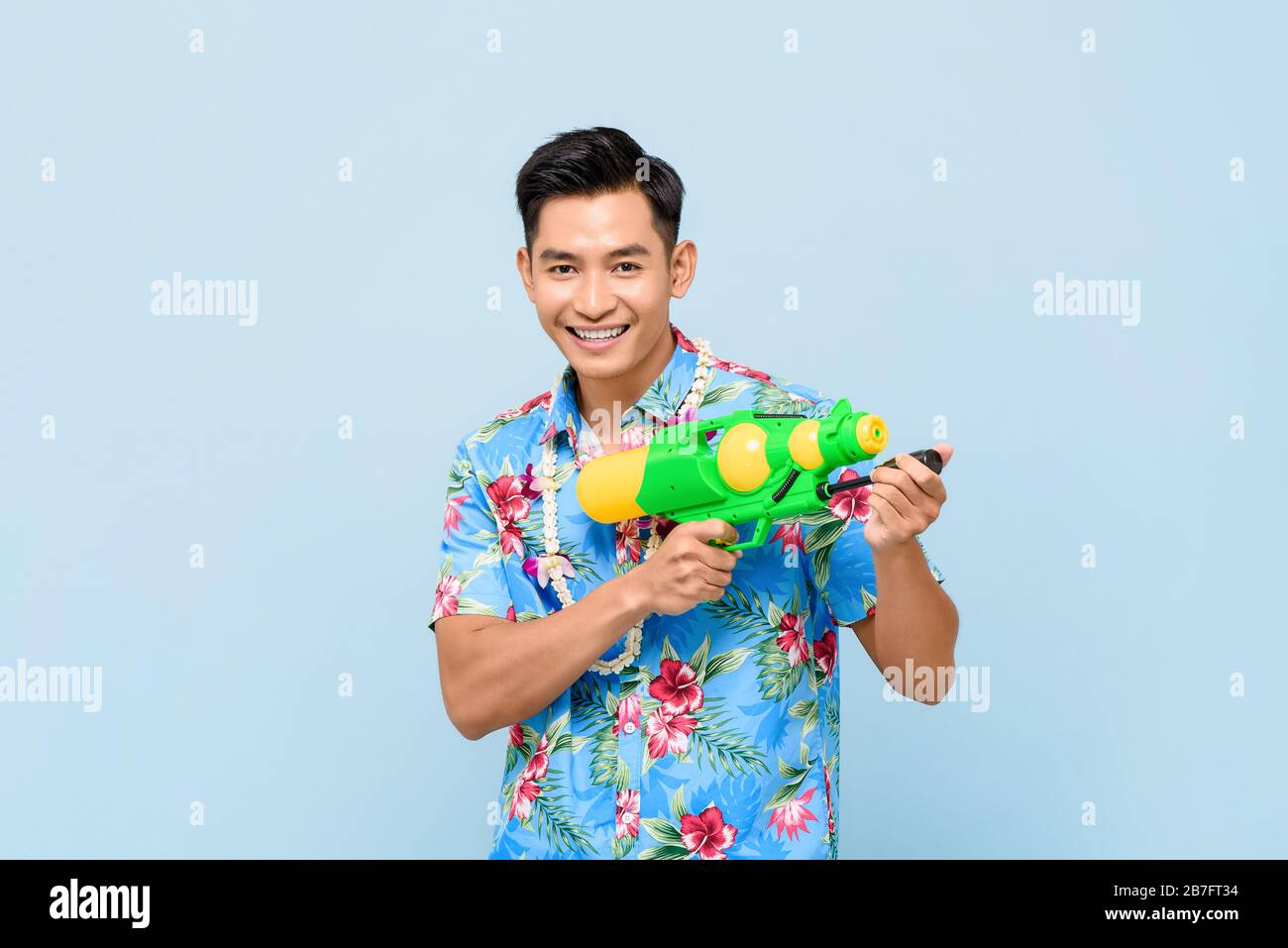Sorridente bell'uomo asiatico che gioca con pistola ad acqua isolato su sfondo blu studio per Songkran festival in Thailandia e sud-est asiatico Foto Stock