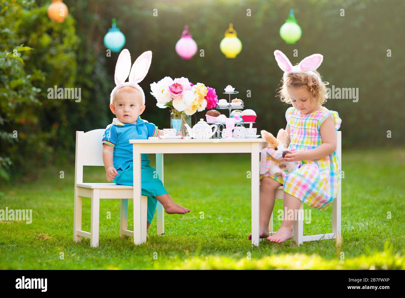 I bambini a caccia di uova di Pasqua. Bambino e ragazza che indossano le orecchie conigliate che giocano la festa del tè mangiando le uova di cioccolato e la torta il giorno di primavera soleggiato. Tradizione familiare Foto Stock