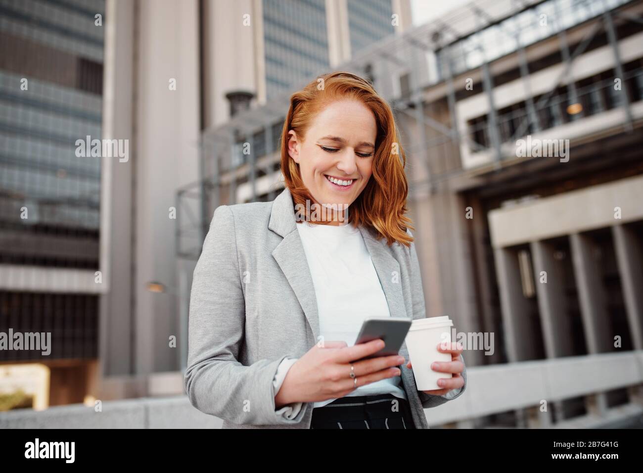 Giovane donna di successo che tiene una tazza da caffè da asporto e utilizza lo smartphone mentre si trova all'aperto Foto Stock