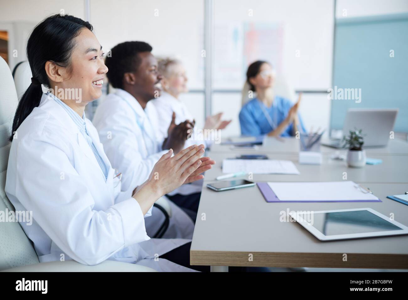 Vista laterale al gruppo multi-etnico di medici applauding mentre siede al tavolo durante la conferenza medica, concentrarsi sul sorridente donna asiatica in primo piano, co Foto Stock