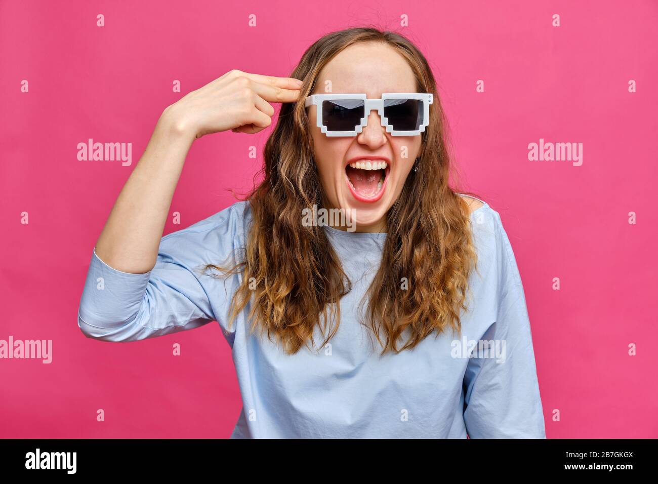 Elegante ragazza caucasica in una t-shirt blu pallido e occhiali a 8 bit e una pistola astratta dalla sua mano spinto alla testa sorridente su uno sfondo rosa. Clo Foto Stock