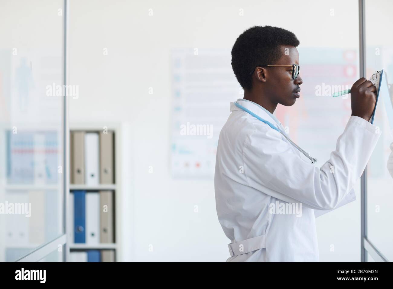 Vista laterale ritratto di giovane medico afro-americano iscritto sulla clipboard mentre in piedi da parete di vetro in clinica medica, spazio copia Foto Stock