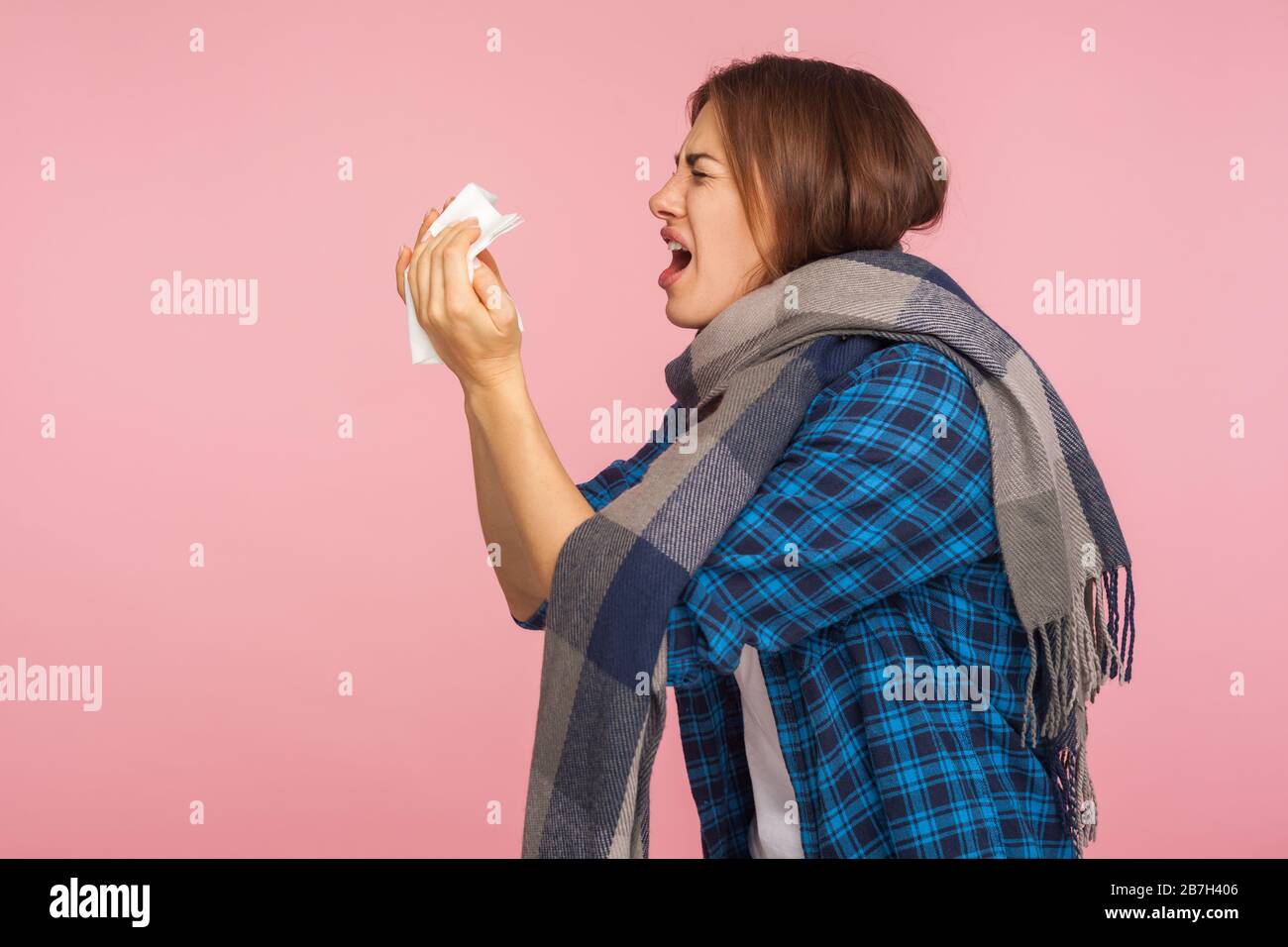 Influenza stagionale. Vista laterale della ragazza malata in camicia a scacchi avvolta in sciarpa che soffia naso runny, starnuti in tovkin, sofferenza infezione virale sintomi. i Foto Stock