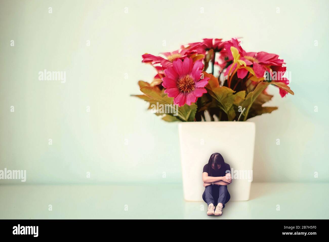 Triste donna depressa seduta con la schiena appoggiata contro bianco vaso con falso fiore rosso. Concetto minimo di tristezza, solitudine. Foto Stock