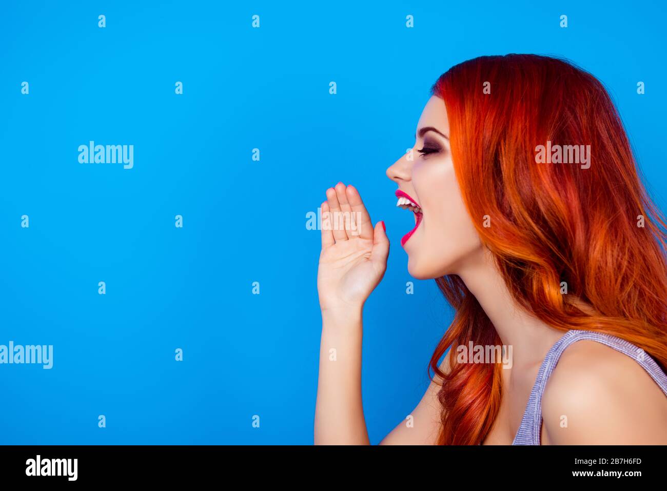 Vista laterale foto di bella ragazza attraente raccontare le notizie mentre la bocca aperta e tenendo le mani vicino alle labbra mentre in piedi su sfondo blu Foto Stock