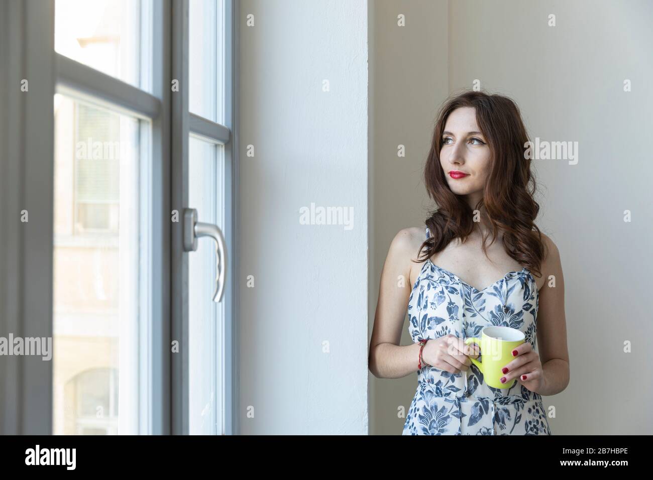 Giovane ragazza rimane alla finestra in piedi con una tazza nella sua mano pensierosamente Foto Stock