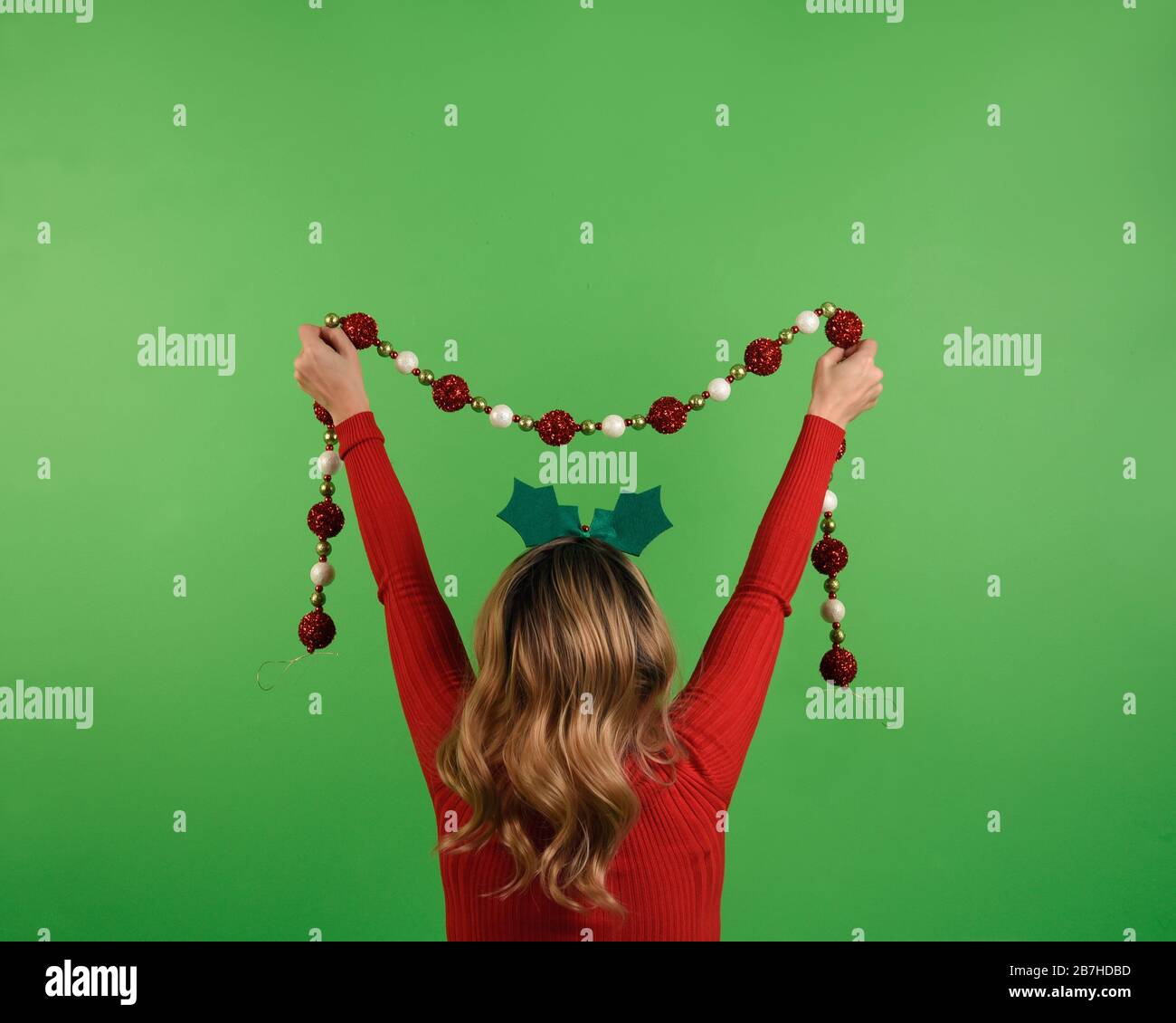 Una donna è in piedi contro una parete verde vuota con copyspace che tiene una decorazione festosa del garland della stringa. Foto Stock