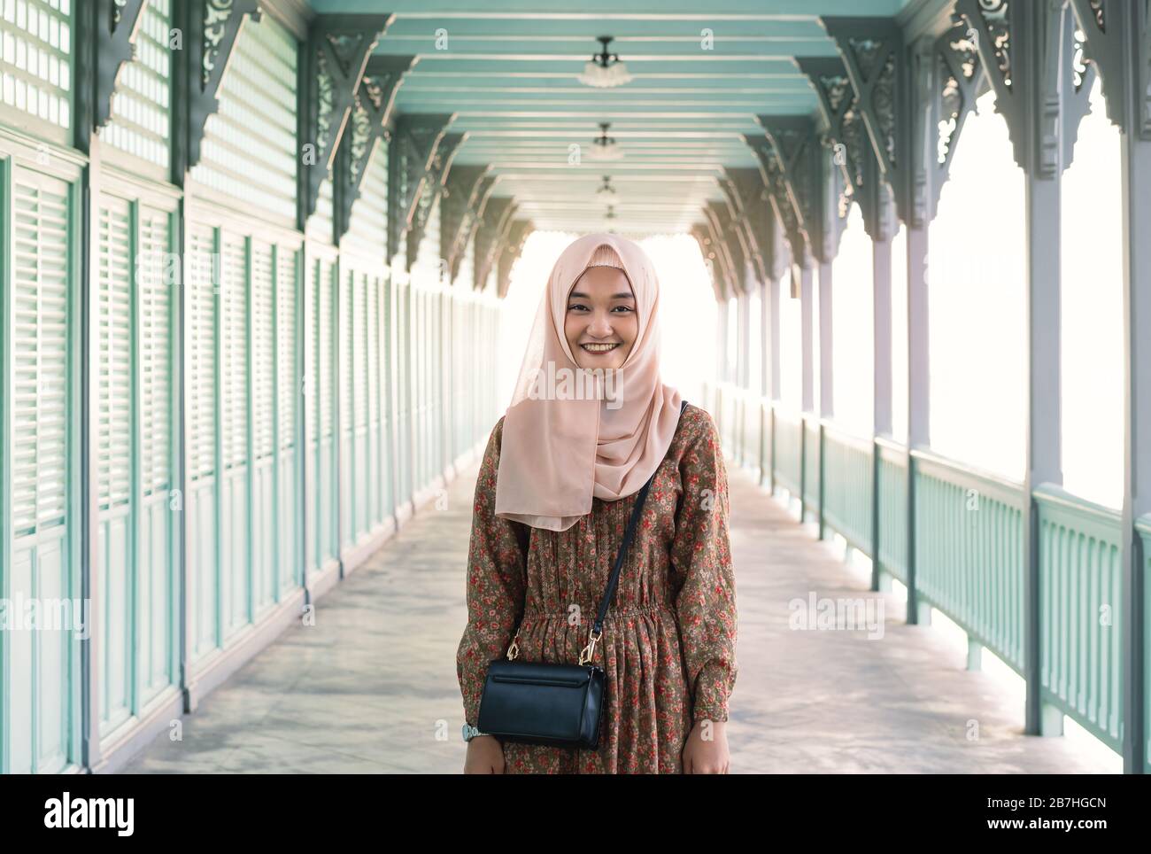 Giovane, felice ragazza musulmana asiatica in piedi all'aperto con sole del pomeriggio luminoso dietro di lei - donna sorridente millenario che indossa il headsharf religioso e abito floreale - Cultura e concetto di diversità Foto Stock