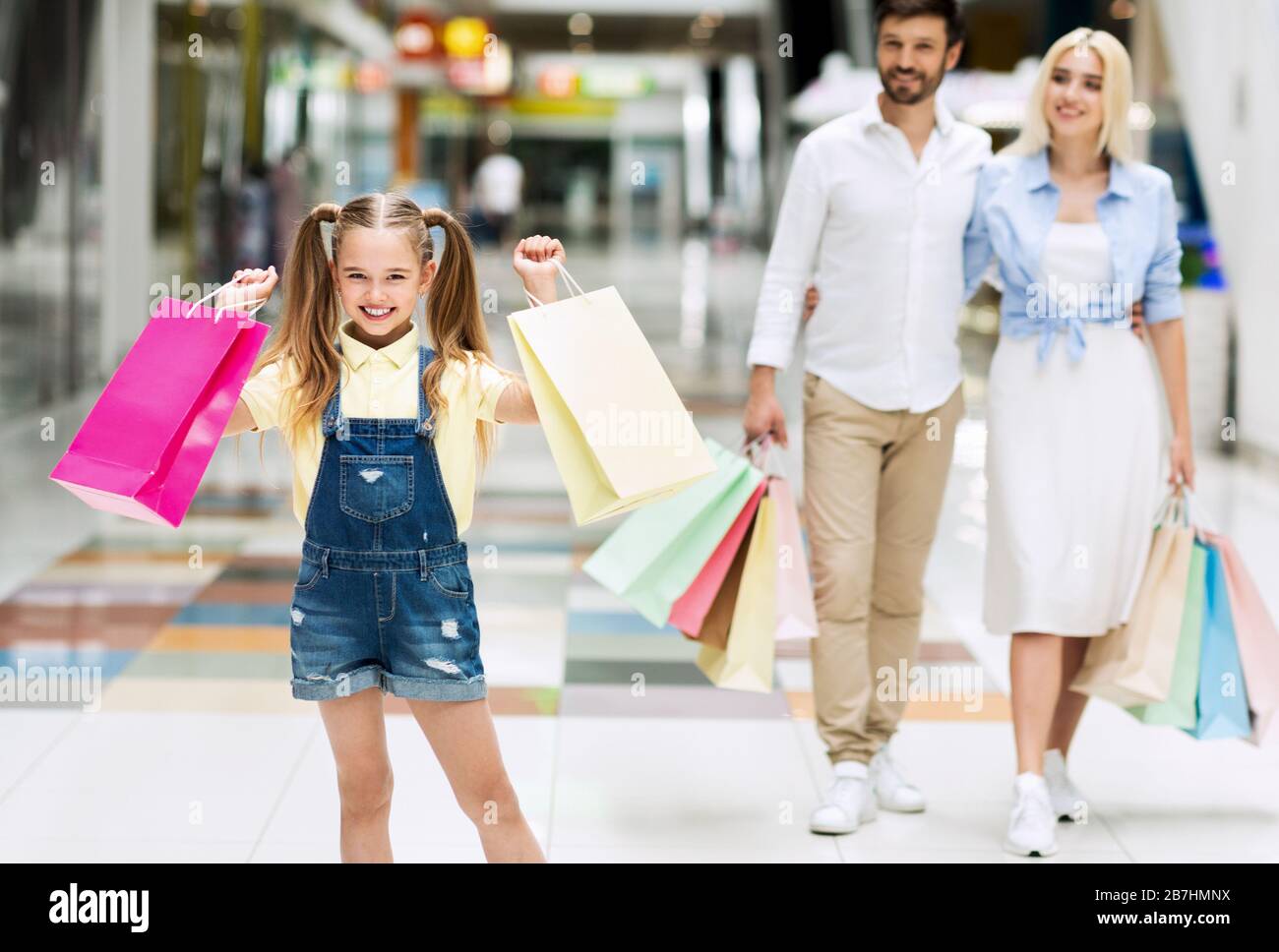 Ragazza che trasporta le borse che comprano i vestiti con i genitori nel centro commerciale Foto Stock
