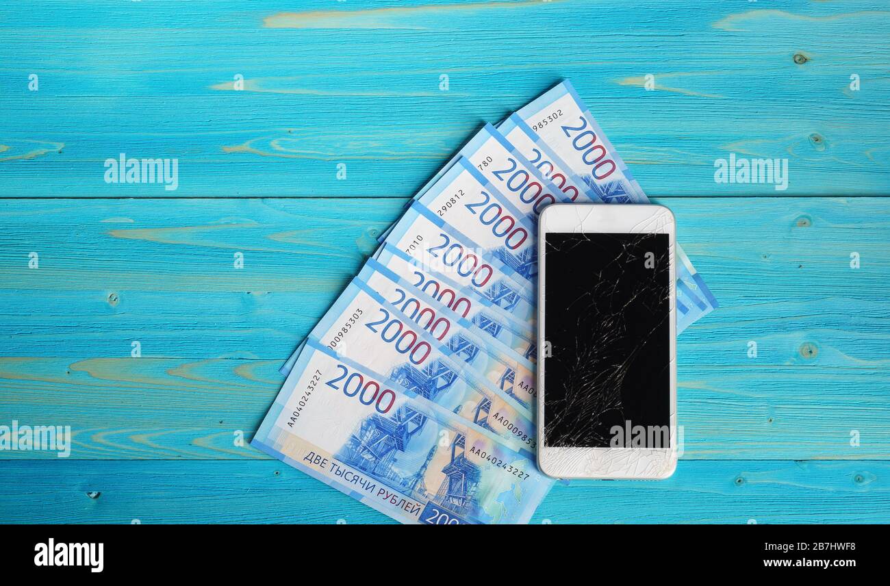 Smartphone con uno schermo rotto e diciottomila rubli nuove bollette sono su un tavolo di legno blu. Costi dei nuovi gadget Foto Stock