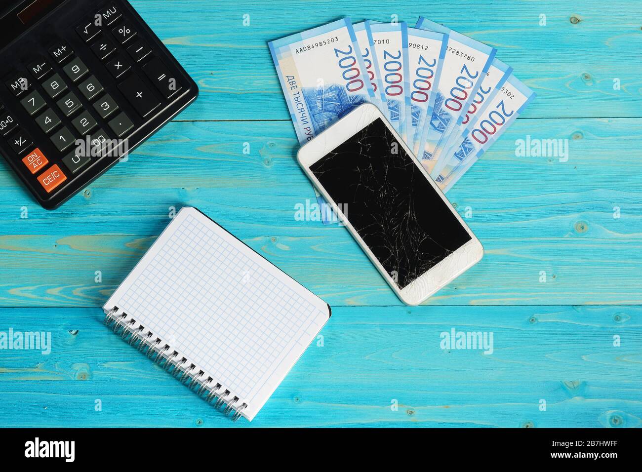 Notebook, smartphone con schermo rotto e quattordici migliaia di rubli nuove fatture giacciono su un tavolo di legno blu. Costi dei nuovi gadget Foto Stock