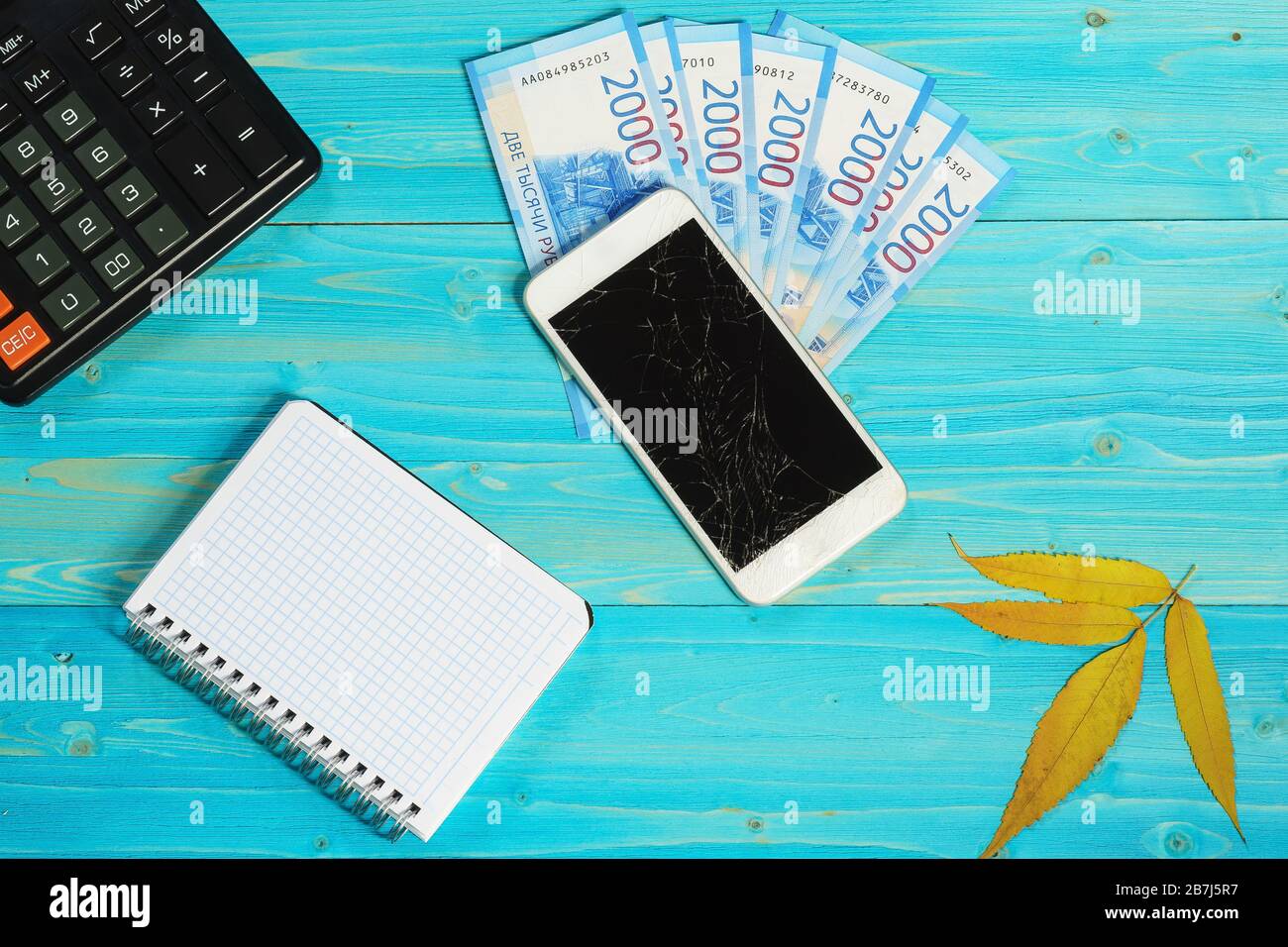 Un notebook, uno smartphone con uno schermo rotto e nuove banconote russe giacciono su un tavolo di legno blu. Il costo di acquisto del gadget Foto Stock