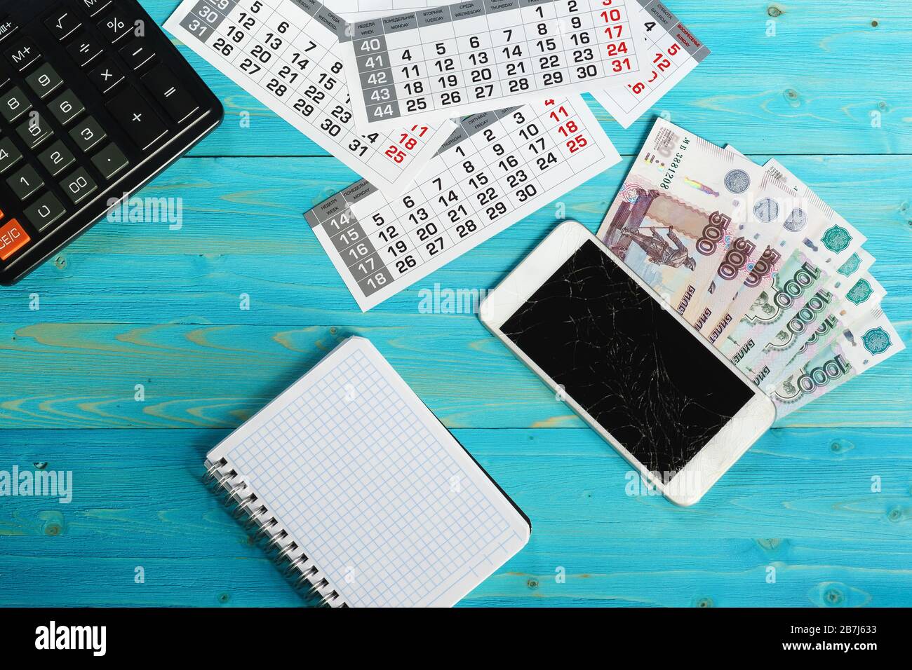 Calendario, smartphone rotto e denaro russo su un tavolo di legno blu. Le spese impreviste hanno interrotto i piani Foto Stock