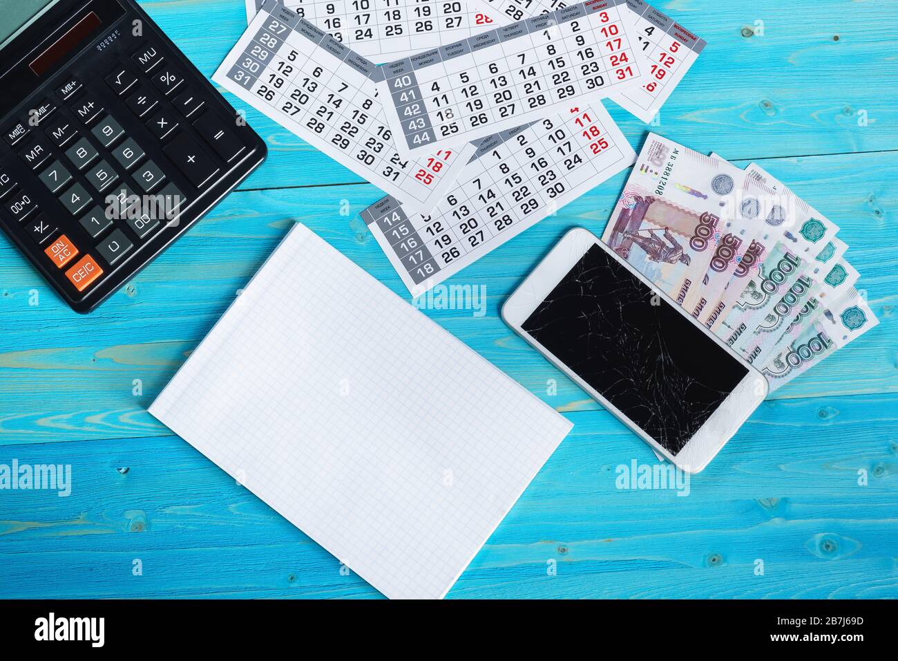 Calendario, notebook pulito, smartphone rotto e denaro russo su un tavolo di legno blu. Le spese impreviste hanno interrotto i piani Foto Stock