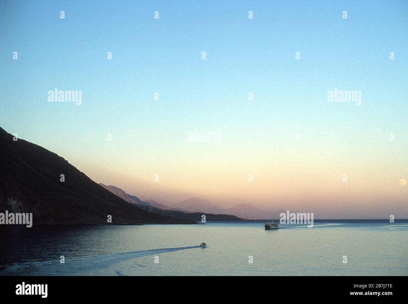 Il traghetto Daskalogiannis su un tranquillo Mar Libico che si avvicina al villaggio di Loutro, Creta, Grecia al tramonto, con la luna piena nel cielo e le montagne dietro. Un motoscafo si dirige in direzione opposta, verso Chora Sfakia, visto sullo sfondo. Foto Stock
