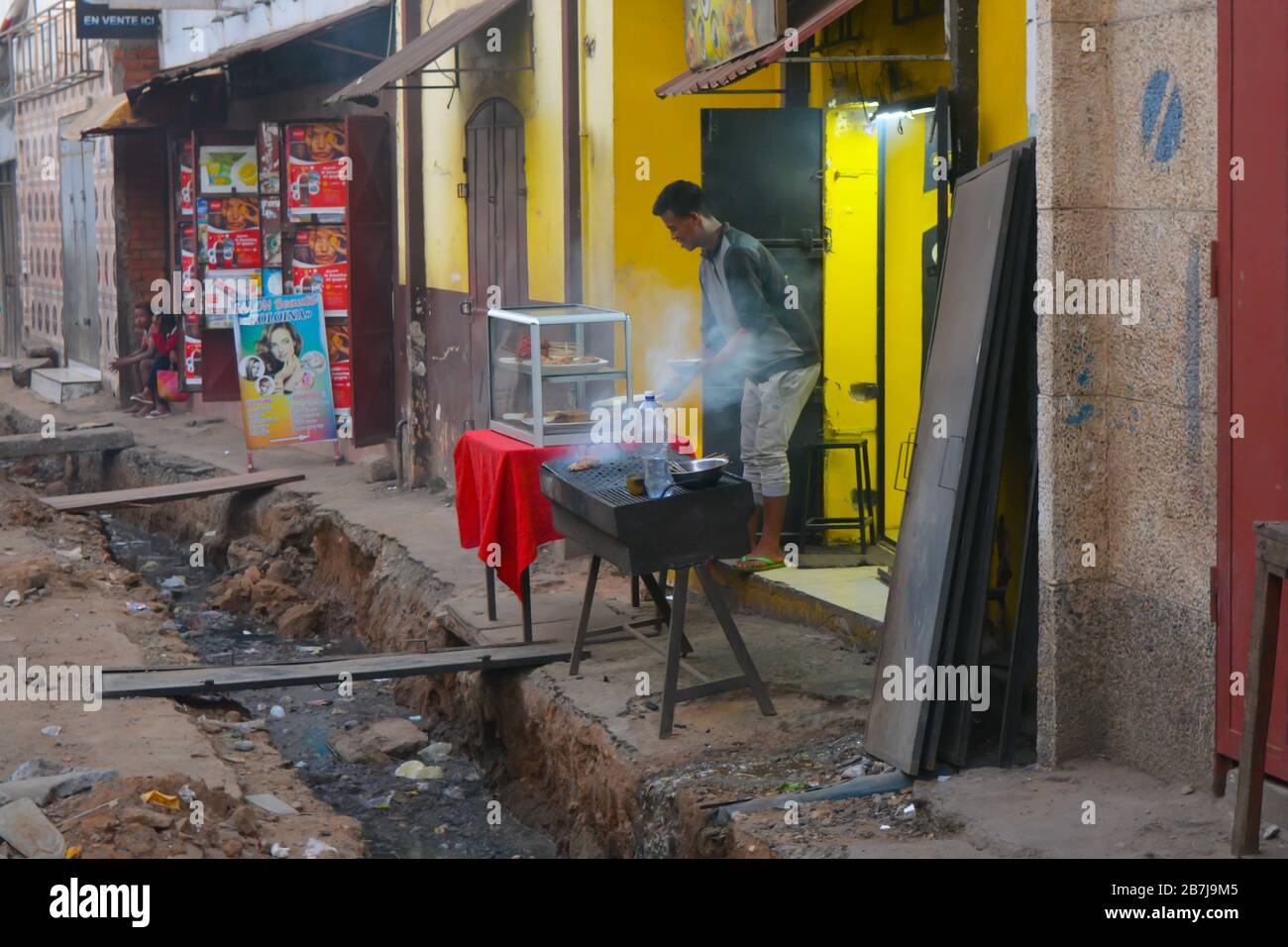 Un uomo africano cuoce con una griglia da fumo sopra un sistema di liquame aperto. Grande buco in strada acqua sporca che scorre. Ristorante malgascio ad Antananarivo Foto Stock