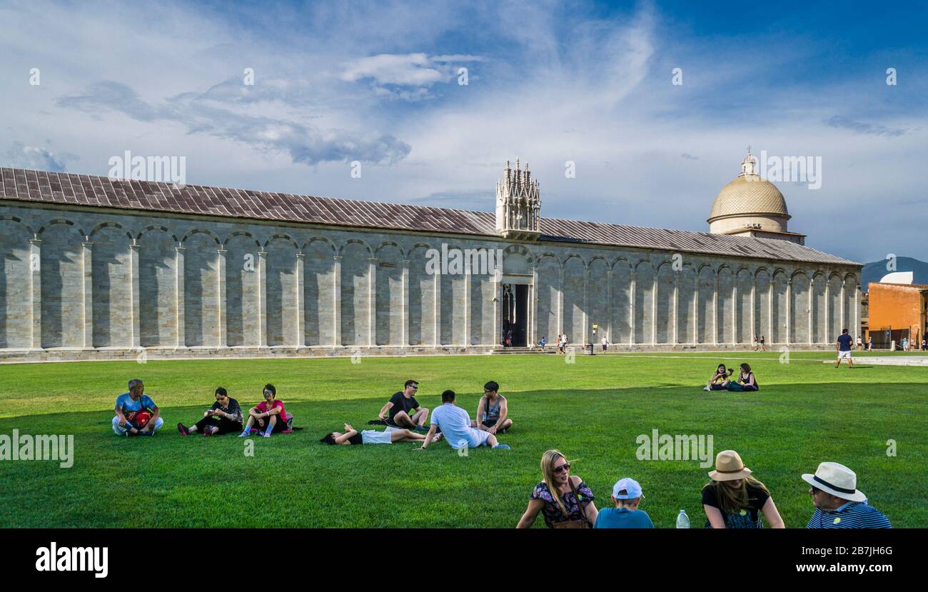 I turisti si riposano sui prati di Piazza dei Miracoli sullo sfondo della parete esterna del Camposanto Monumentale, Pisa, Toscana, Foto Stock