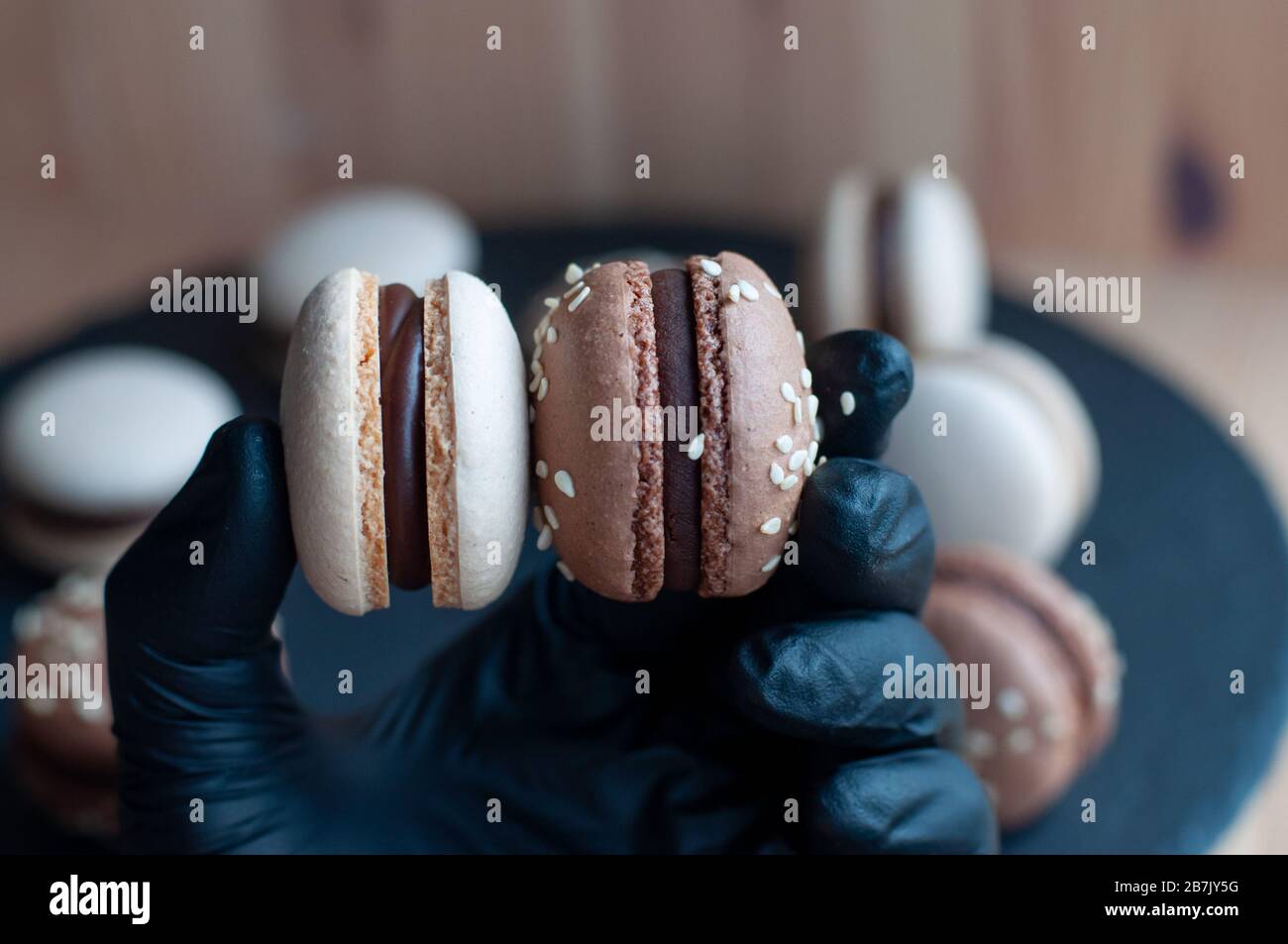 Mano in guanti neri per due macaron con ganache al cioccolato e semi di sesamo Foto Stock